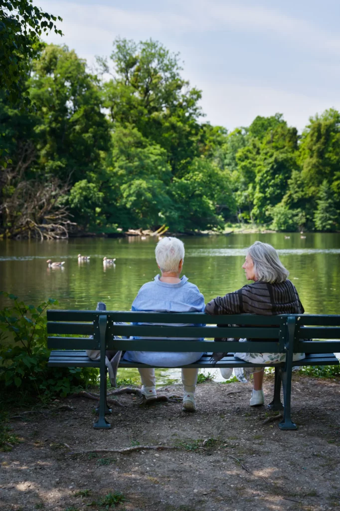 Zwei Personen sitzen auf einer Parkbank an einem Teich, unterhalten sich. Enten schwimmen auf dem Teich. Die Personen sind von hinten fotografiert