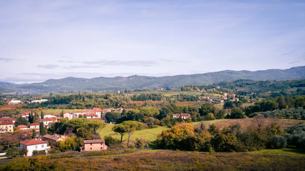 Eine Landschaft in der Toskana mit rollenden Hügeln, einigen Häusern, Zypressen, Olivenbäumen und einem Gebirge im Hintergrund. Der Himmel ist leicht bewölkt und blau