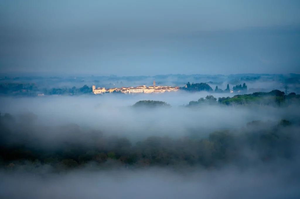 Leuchtendes Dorf in der Toskana eingehüllt in den herbstlichen Morgennebel. Das Wetter ist gedrückt, dunkel.