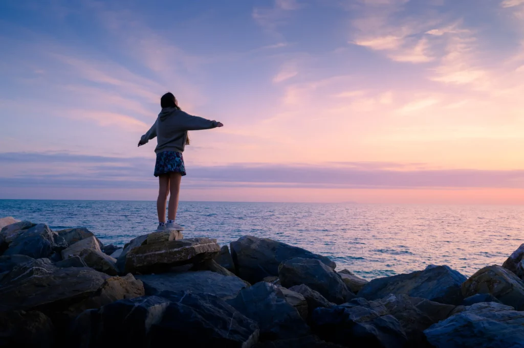 Eine Person steht auf großen Steinen am Meer und schaut in den Sonnenuntergang. Einige Wolken sind am Himmel
