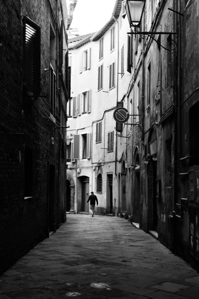 Eine Gasse in Sienna mit hohen Häusern. Eine Person ist im Hintergrund zu sehen.