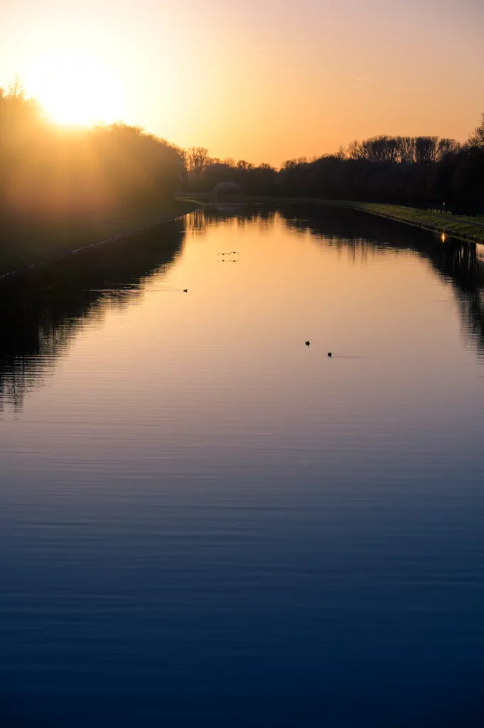 Blick entlang eines ruhigen Kanals bei Sonnenuntergang. Der Himmel leuchtet orange-gelb, die Wasseroberfläche spiegelt das Licht wider. Vereinzelte Wasservögel sind auf dem Wasser zu sehen, am Horizont Silhouetten von Bäumen.